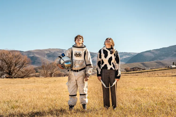 Zdjęcie; Chłopak w stroju kosmonauty i dziewczyna wpatrują się w niebo stojąc w polu