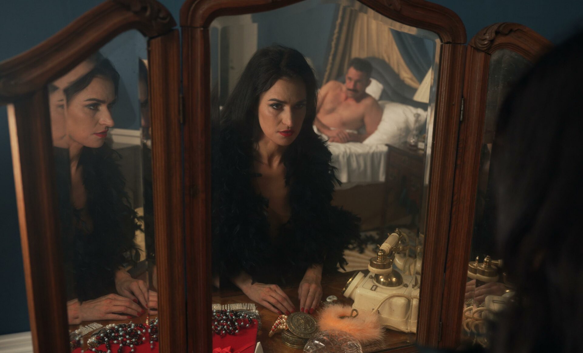 Zdjęcie; Kobieta w mocnym makijażu patrzy w lustro, w którym odbija się nagi mężczyzna na łóżku