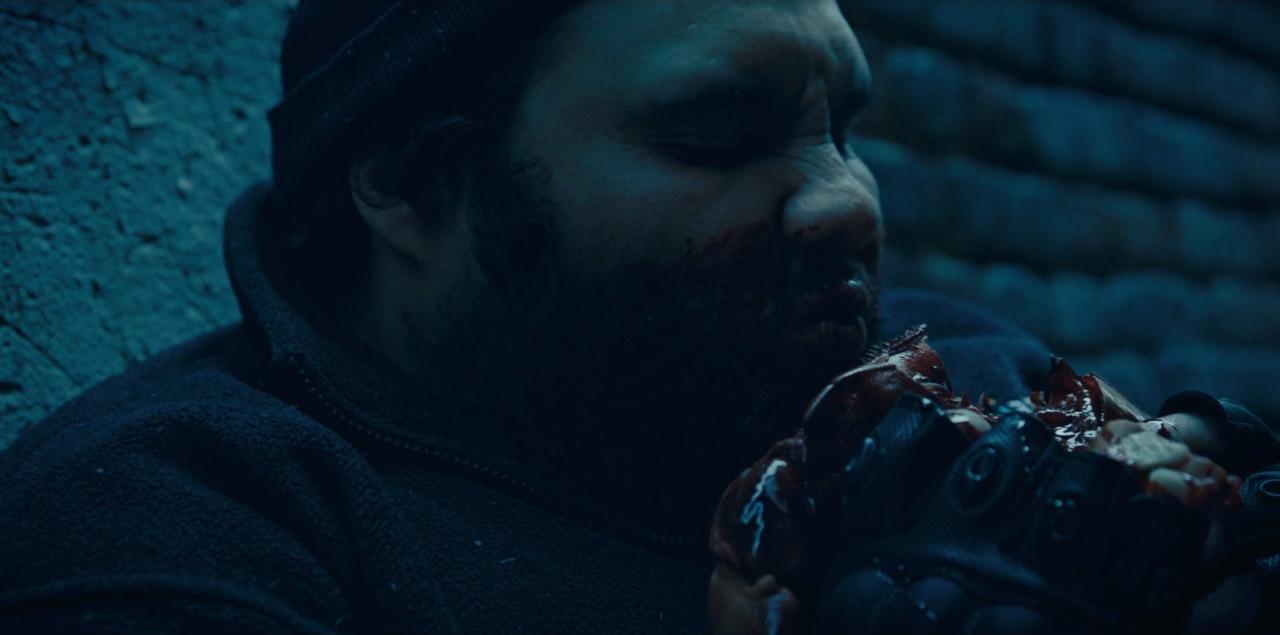 Zdjęcie; Mężczyzna w czarnej bluzie, z twarzą brudną od krwi opiera się o ścianę i trzyma coś zakrwawionego